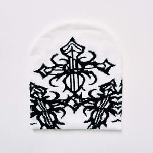 Könnte beinhalten: Weiße Strickmütze mit einem schwarz-weißen Kreuzmuster. Das Kreuz ist mit komplizierten Details und einem tribal inspirierten Muster gestaltet.