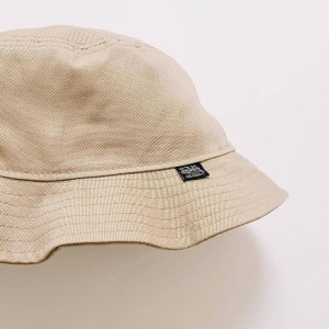 Puede incluir: Un sombrero de pescador beige con una etiqueta negra "Von Dutch Originals" en el lateral.
