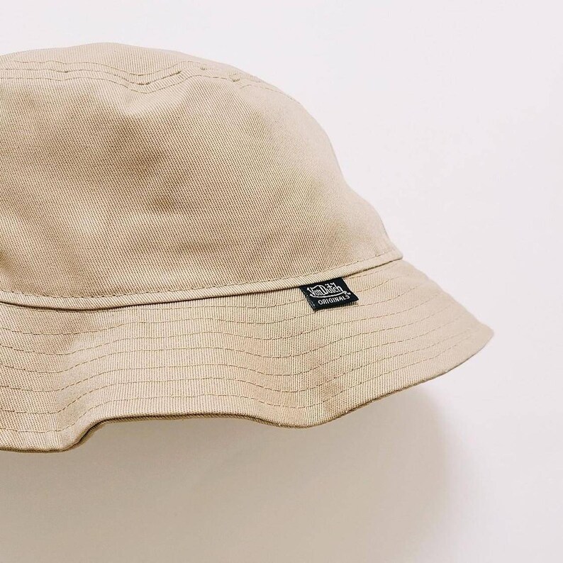 Puede incluir: Un sombrero de pescador beige con una etiqueta negra "Von Dutch Originals" en el lateral.