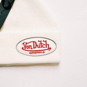 May include: Cream-colored beanie hat with a red and white embroidered Von Dutch Originals logo. The hat is made of a soft, textured material and features a classic, folded cuff design. A black tag is attached to the hat.