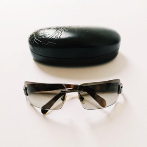 May include: A pair of brown and silver sunglasses with a rimless design. The sunglasses are sitting on a white surface. The sunglasses come with a black case.