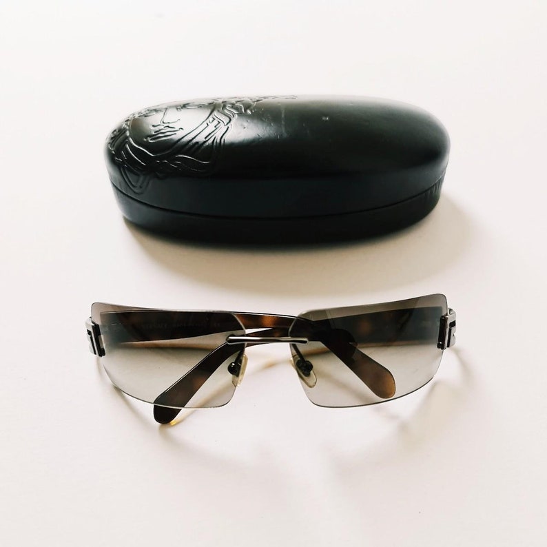 May include: A pair of brown and silver sunglasses with a rimless design. The sunglasses are sitting on a white surface. The sunglasses come with a black case.