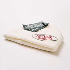 May include: Cream-colored knit beanie with a red and silver Von Dutch patch. A black tag with the Von Dutch logo is attached. The beanie is folded and lies on a white surface. The beanie is a casual accessory.