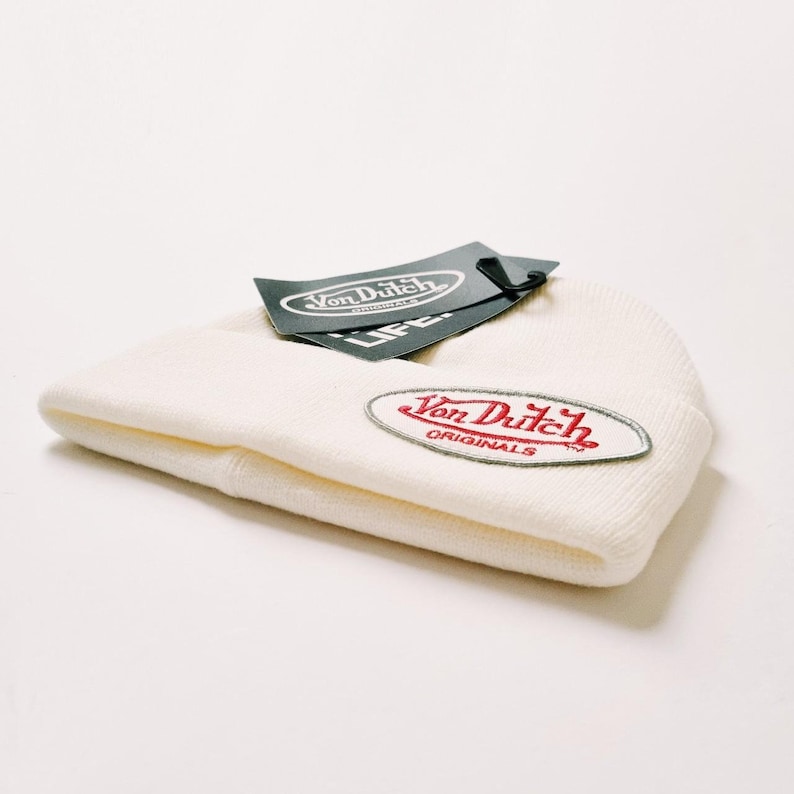 May include: Cream-colored knit beanie with a red and silver Von Dutch patch. A black tag with the Von Dutch logo is attached. The beanie is folded and lies on a white surface. The beanie is a casual accessory.