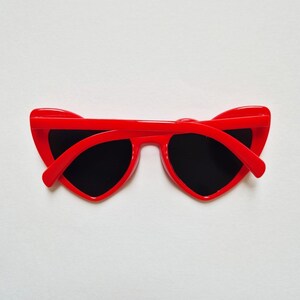 May include: Red heart-shaped sunglasses with black lenses. The sunglasses are folded in half and lying on a white surface.