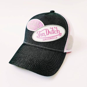 Puede incluir: Una gorra de camionero negra y blanca con un parche de logotipo rosa "Von Dutch Originals". La gorra tiene una parte trasera de malla.