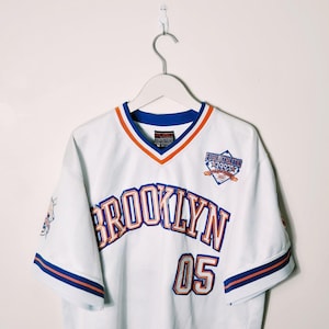 Può includere: Maglia da baseball bianca con bordini blu e arancioni. La maglia ha "BROOKLYN" scritto in blu e arancione sul davanti con il numero "05" sotto. La maglia ha anche una toppa sul lato destro che dice "FUBU ATHLETICS 1992".