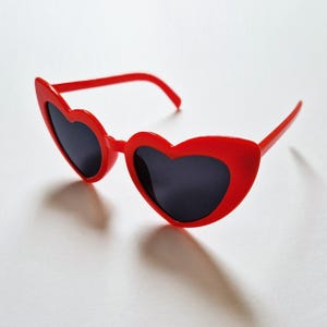 May include: Red heart-shaped sunglasses with black lenses. The sunglasses are made of plastic and have a simple design.