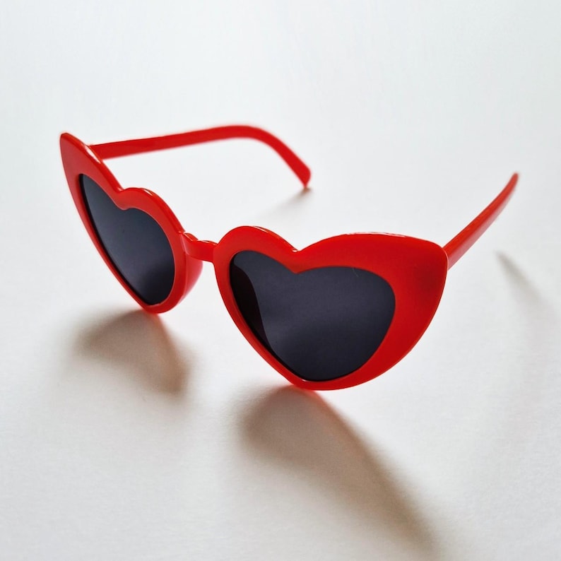 May include: Red heart-shaped sunglasses with black lenses. The sunglasses are made of plastic and have a simple design.