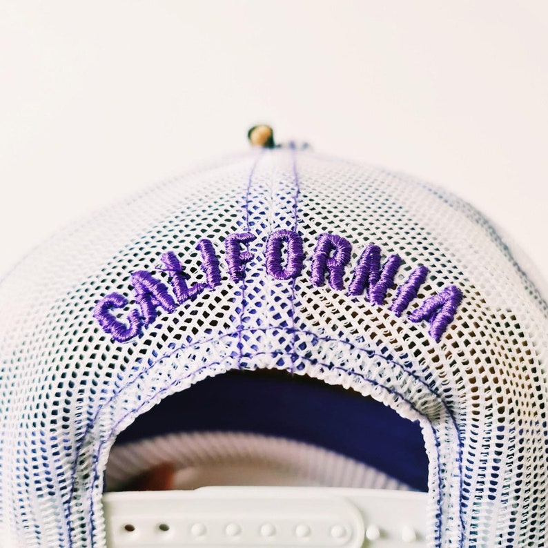 May include: White mesh-backed baseball cap with the word "CALIFORNIA" embroidered in purple across the back. The cap has a white adjustable strap and a purple interior.