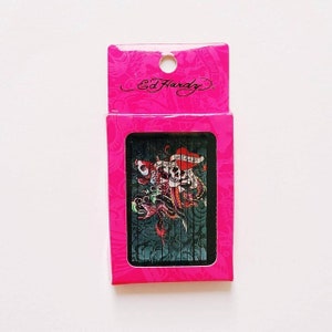 K&ouml;nnte beinhalten: Eine rosa Box mit den Worten "Ed Hardy" auf der Vorderseite. Die Box enth&auml;lt ein Kartenspiel mit einem Totenkopf- und Schlangenmotiv. Die Karten haben einen schwarzen Hintergrund mit einem roten Herzen und den Worten "Love Kills" auf der Vorderseite.
