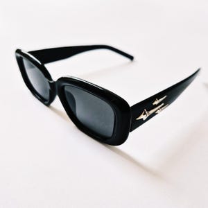 K&ouml;nnte beinhalten: Schwarze rechteckige Sonnenbrille mit silbernem Akzent an der Seite. Die Sonnenbrille hat dunkle Gl&auml;ser.