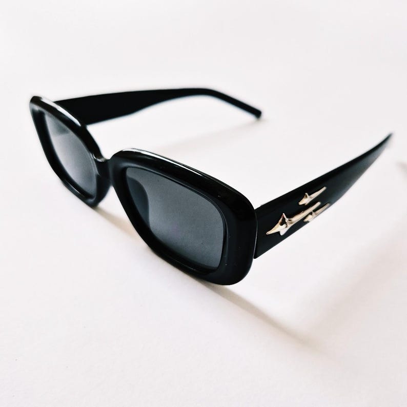 K&ouml;nnte beinhalten: Schwarze rechteckige Sonnenbrille mit silbernem Akzent an der Seite. Die Sonnenbrille hat dunkle Gl&auml;ser.