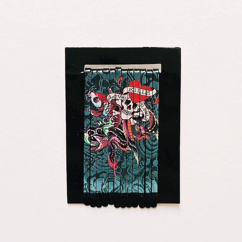 K&ouml;nnte beinhalten: Ein Satz schwarzer Haarklammern mit einem bunten Design, das einen Totenkopf, ein Herz und ein Banner mit der Aufschrift "Love Kills Slowly" zeigt.