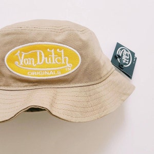 Puede incluir: Un sombrero de pescador beige con un parche ovalado amarillo que dice "Von Dutch Originals" en letras blancas. El sombrero tiene una etiqueta negra adjunta.