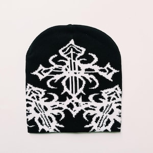 Könnte beinhalten: Schwarze Strickmütze mit einem weißen Kreuzmuster. Das Kreuz ist von einem weißen Blumenmuster umgeben.