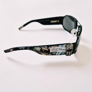 Może przedstawiać: Czarne okulary przeciwsłoneczne z kolorowym wzorem Ed Hardy na zausznikach. Okulary mają czarną oprawkę i ciemnoszare szkła. Wz&oacute;r na zausznikach przedstawia węża, kostki do gry i podpis Ed Hardy.