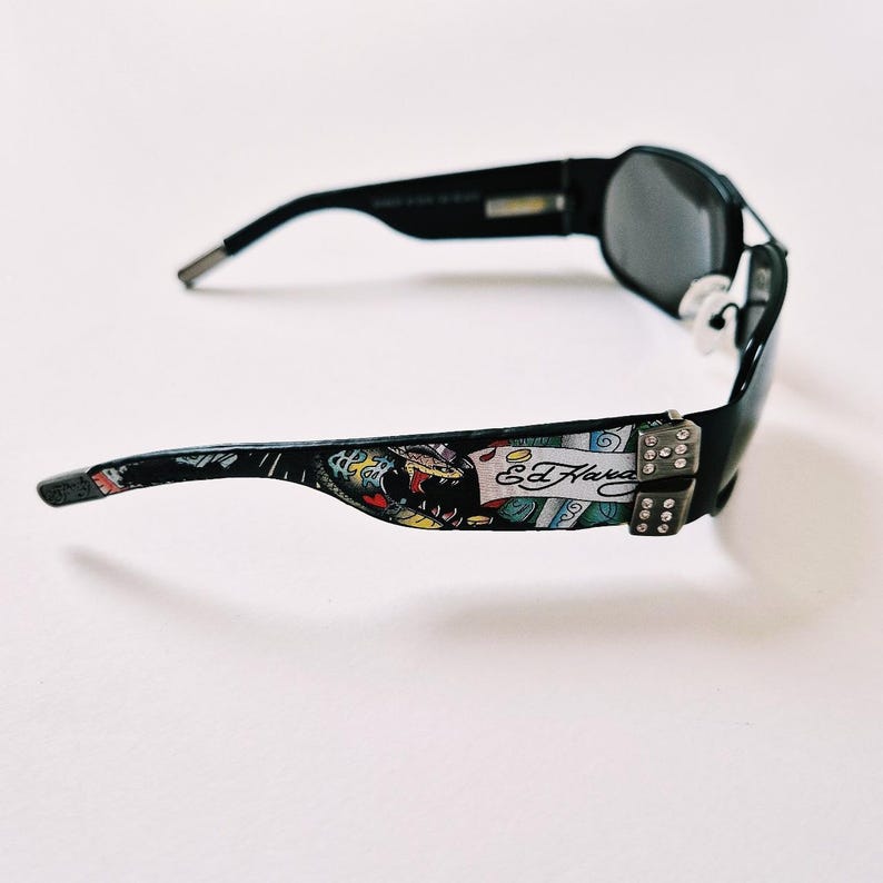 Może przedstawiać: Czarne okulary przeciwsłoneczne z kolorowym wzorem Ed Hardy na zausznikach. Okulary mają czarną oprawkę i ciemnoszare szkła. Wz&oacute;r na zausznikach przedstawia węża, kostki do gry i podpis Ed Hardy.