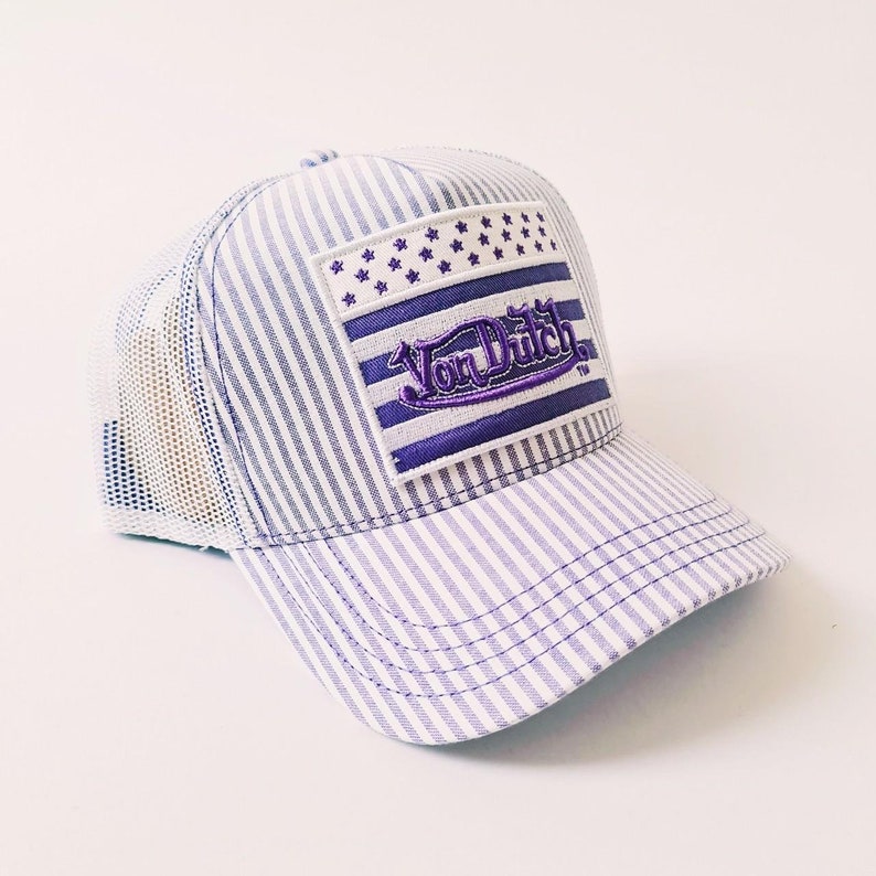 May include: A baseball cap with a striped pattern and a mesh back. The cap has a purple and white striped design and a rectangular patch with the text "Von Dutch" in purple. The patch also features a stars and stripes design.