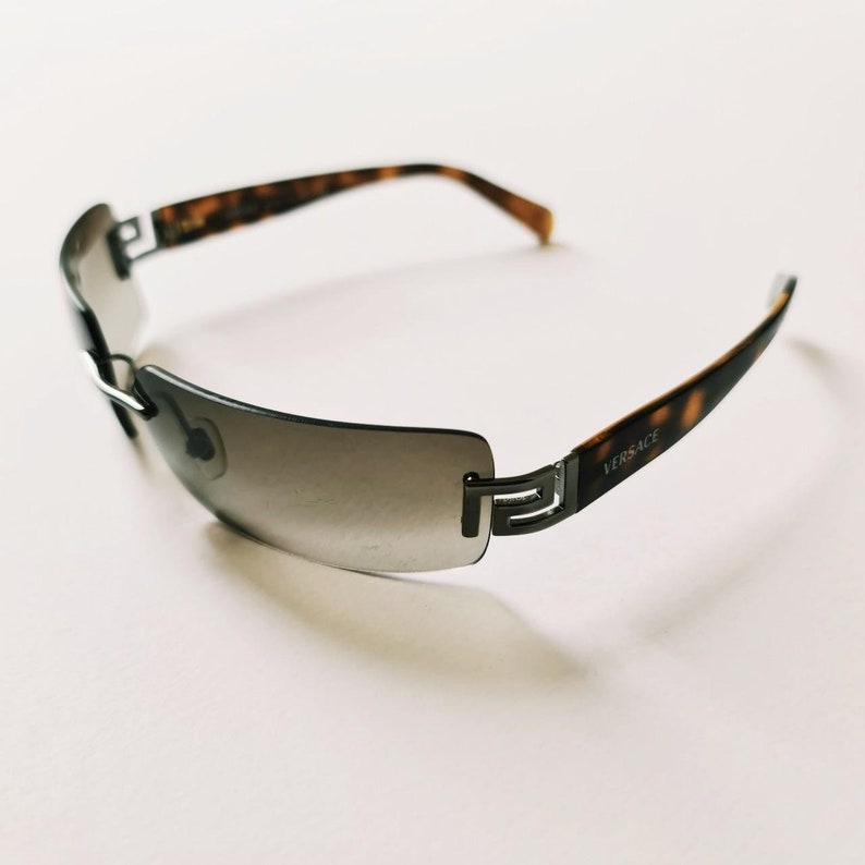 May include: A pair of brown and silver sunglasses with a gradient grey lens. The sunglasses have a Versace logo on the side.