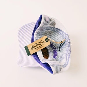 May include: A Von Dutch baseball cap with a striped front panel and a white mesh back. The cap features a purple brim and the Von Dutch California logo on a brown tag.