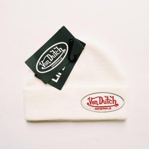 May include: Cream-colored beanie with a red and white Von Dutch Originals logo patch. A dark green tag with the Von Dutch logo hangs from the beanie. The beanie is made of a soft, knitted material.