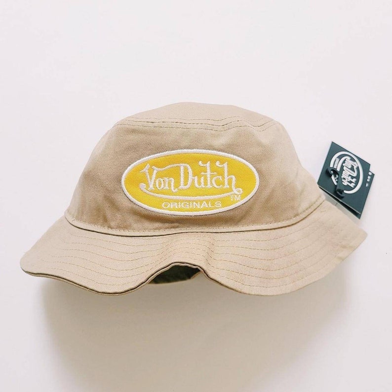 Puede incluir: Un sombrero de pescador beige con un parche ovalado amarillo que dice "Von Dutch Originals TM" en letras blancas.