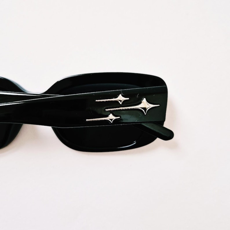 K&ouml;nnte beinhalten: Schwarze rechteckige Sonnenbrille mit einem silbernen Sternendesign auf dem Rahmen.