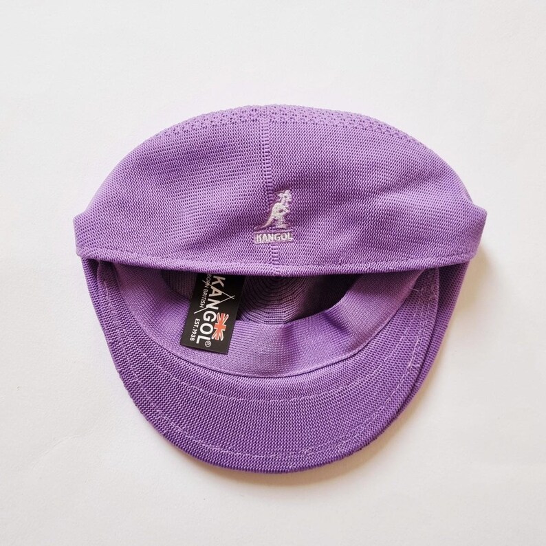 May include: A purple knitted Kangol brand baseball cap with a kangaroo logo on the front. The cap has a small label on the side that says "Kangol, Made in Britain, Est 1938."