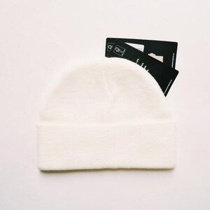 May include: A cream-colored beanie hat with a folded brim. The hat is made of a soft, knitted material. Two black tags are partially visible behind the hat. The tags have white text and a small graphic.