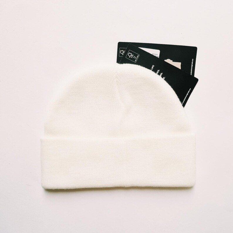 May include: A cream-colored beanie hat with a folded brim. The hat is made of a soft, knitted material. Two black tags are partially visible behind the hat. The tags have white text and a small graphic.