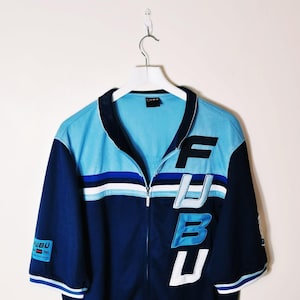 Può includere: Giacca blu e navy con zip con la scritta "FUBU" a grandi lettere bianche e nere sul davanti. La giacca ha una sezione superiore azzurra e un design a righe bianche. Le maniche sono blu navy.