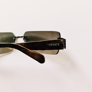 May include: Brown tortoiseshell sunglasses with a rectangular frame and brown lenses. The sunglasses have a metal bar connecting the two lenses and the brand name "VERSACE" is printed on the right arm.