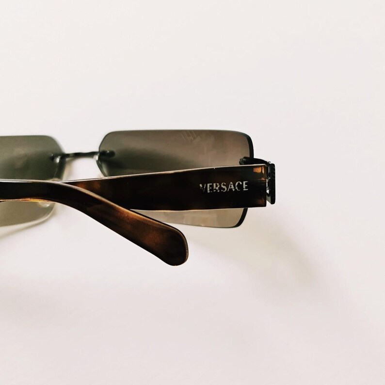 May include: Brown tortoiseshell sunglasses with a rectangular frame and brown lenses. The sunglasses have a metal bar connecting the two lenses and the brand name "VERSACE" is printed on the right arm.