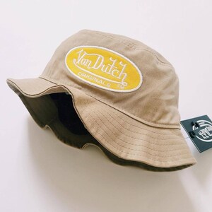 Puede incluir: Un sombrero de pescador beige con un parche ovalado amarillo que dice "Von Dutch Originals TM" en letras blancas.