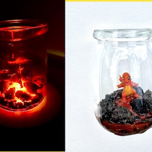 Puede incluir: Un frasco de vidrio transparente con un brillo rojo desde adentro, lleno de lo que parecen brasas brillantes. El frasco está sobre una superficie blanca. Un segundo frasco contiene una pequeña figura naranja y amarilla sobre un lecho de rocas oscuras.