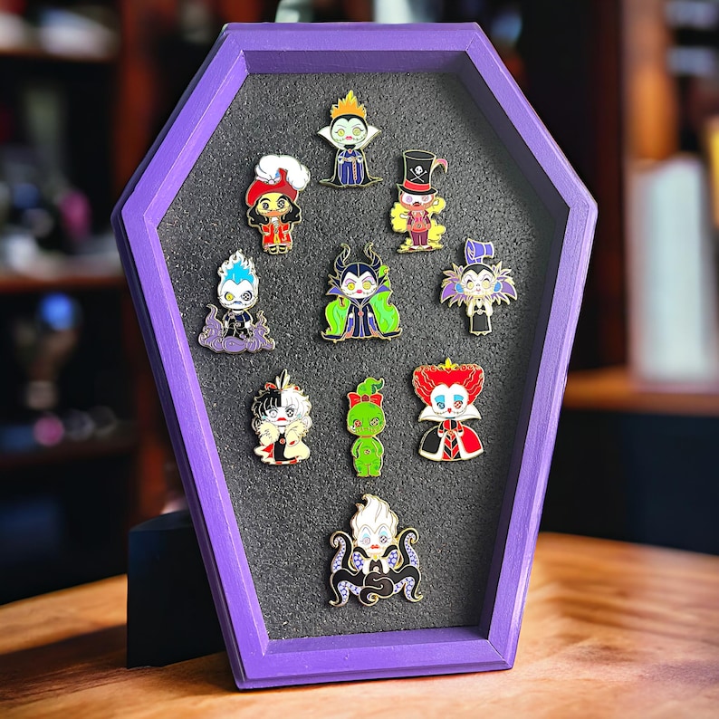 Pode incluir: Uma cole&ccedil;&atilde;o de pins de esmalte com v&aacute;rios vil&otilde;es de desenhos animados, exibidos numa caixa de sombra roxa em forma de caix&atilde;o. Os pins apresentam personagens em cores vibrantes, com designs detalhados.