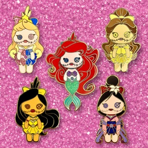 Pode incluir: Cinco pinos de esmalte com personagens estilizados de bonecas com olhos de bot&atilde;o e bocas costuradas. Os personagens incluem uma sereia com cabelo vermelho, uma princesa com um vestido amarelo e outras figuras com roupas coloridas. Os pinos t&ecirc;m contornos dourados.