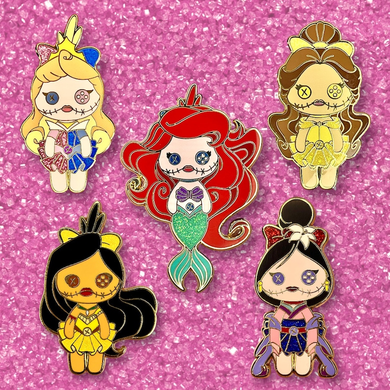 Pode incluir: Cinco pinos de esmalte com personagens estilizados de bonecas com olhos de bot&atilde;o e bocas costuradas. Os personagens incluem uma sereia com cabelo vermelho, uma princesa com um vestido amarelo e outras figuras com roupas coloridas. Os pinos t&ecirc;m contornos dourados.