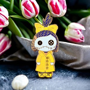 Pode incluir: Um alfinete de esmalte amarelo em forma de boneca com olhos de bot&atilde;o e boca costurada. A boneca usa um casaco amarelo e um la&ccedil;o azul e amarelo. O alfinete &eacute; colocado em um fundo de tulipas rosa e brancas.