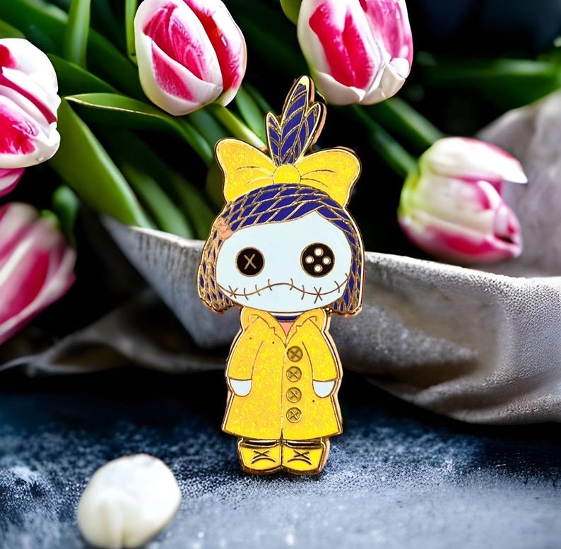 Pode incluir: Um alfinete de esmalte amarelo em forma de boneca com olhos de bot&atilde;o e boca costurada. A boneca usa um casaco amarelo e um la&ccedil;o azul e amarelo. O alfinete &eacute; colocado em um fundo de tulipas rosa e brancas.