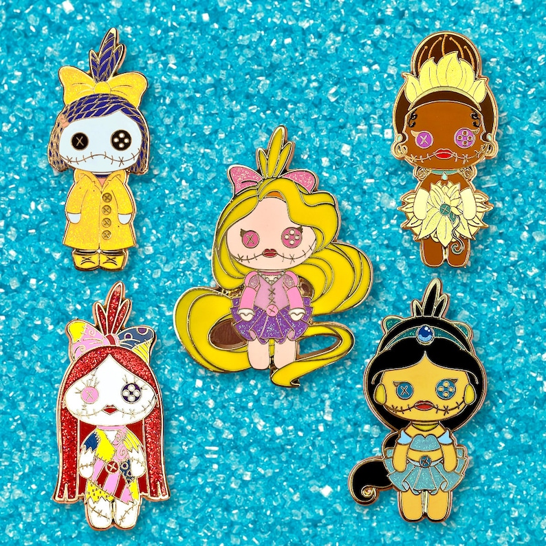 Pode incluir: Pins de esmalte com personagens estilizados semelhantes a bonecas com tra&ccedil;os faciais costurados. Os pins apresentam personagens em v&aacute;rias roupas, incluindo um casaco amarelo, um vestido roxo e uma roupa azul, com contornos dourados. Fundo azul texturizado.