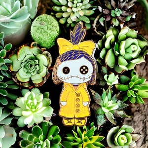 Pode incluir: Um alfinete de esmalte amarelo com um personagem semelhante a uma boneca com olhos de bot&atilde;o, boca costurada e um la&ccedil;o azul e amarelo. O personagem usa um casaco amarelo, rodeado por v&aacute;rias suculentas verdes.