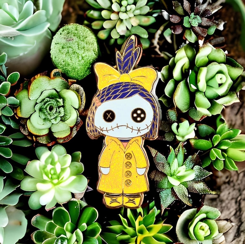 Pode incluir: Um alfinete de esmalte amarelo com um personagem semelhante a uma boneca com olhos de bot&atilde;o, boca costurada e um la&ccedil;o azul e amarelo. O personagem usa um casaco amarelo, rodeado por v&aacute;rias suculentas verdes.