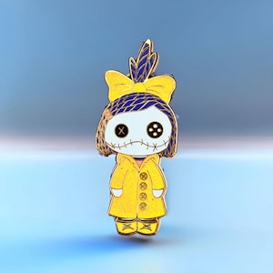 Pode incluir: Um alfinete de esmalte amarelo com um personagem semelhante a uma boneca com boca costurada e olhos de bot&atilde;o. O personagem usa um casaco amarelo e um la&ccedil;o com detalhes azuis. O alfinete tem um contorno dourado em um fundo azul gradiente.