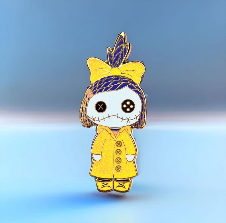 Pode incluir: Um alfinete de esmalte amarelo com um personagem semelhante a uma boneca com boca costurada e olhos de bot&atilde;o. O personagem usa um casaco amarelo e um la&ccedil;o com detalhes azuis. O alfinete tem um contorno dourado em um fundo azul gradiente.
