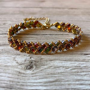 Peut inclure: Un bracelet en perles dorées avec un motif en chevron réalisé avec des perles de verre irisées dans les tons vert, marron et violet.
