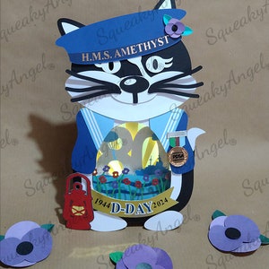 May include: A papercraft model of a cat wearing a blue sailor hat with the text "H.M.S. AMETHYST" on it. The cat is holding a lantern and a medal with the text "PDSA". The cat is standing in front of a scene depicting a beach with a ship in the background. The scene is surrounded by a gold frame with the text "1944 D-DAY 2024". There are three purple poppy flowers on the table.