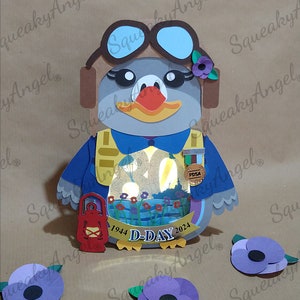 Könnte beinhalten: Eine Papierfigur eines Pinguins, der eine blaue Pilotenuniform und eine Fliegerbrille trägt. Der Pinguin hält eine Laterne und eine Medaille. Die Figur ist mit roten Mohnblumen und dem Text "1944 D-DAY 2024" verziert.