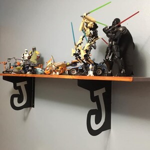 Puede incluir: Una estantería de madera con borde naranja y soportes negros en forma de la letra "J" sostiene varias figuras de Lego de Star Wars. Las figuras incluyen a Darth Vader, el General Grievous y otros personajes con sables de luz.